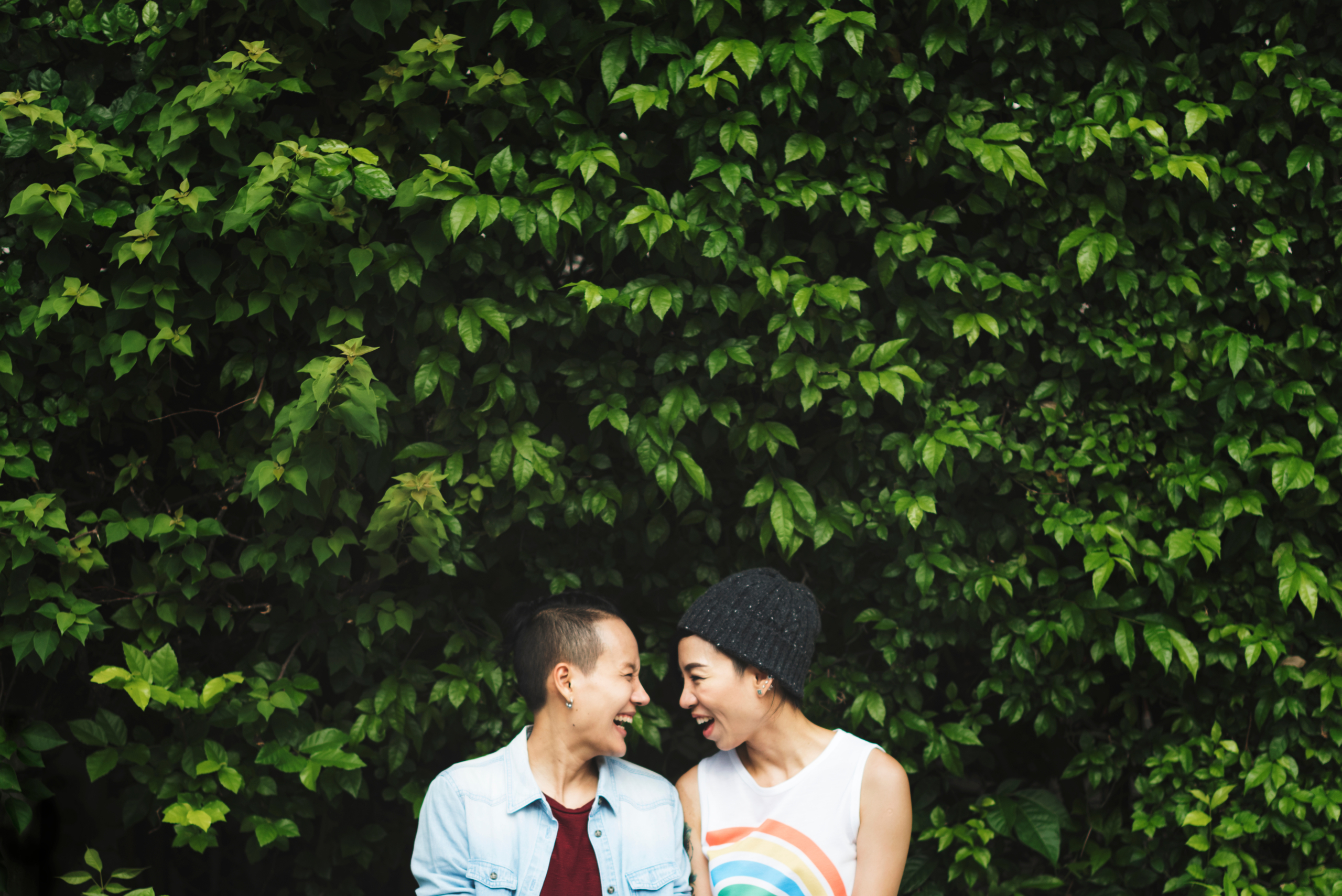 Two Asian people smiling at each other in front of a large bush. One person is wearing a rainbow shirt.